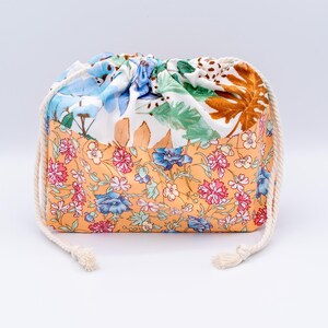 May include: A drawstring pouch featuring a two-tone design. The bottom section displays a peach-coloured fabric with a dense floral pattern in pink, blue, and green. The top section is white with a botanical print of green leaves and brown foliage.