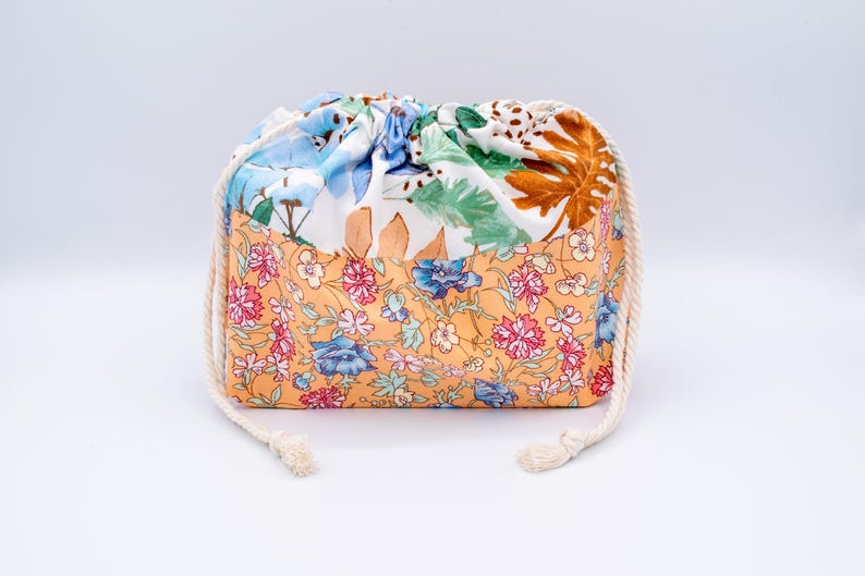 May include: A drawstring pouch featuring a two-tone design. The bottom section displays a peach-coloured fabric with a dense floral pattern in pink, blue, and green. The top section is white with a botanical print of green leaves and brown foliage.