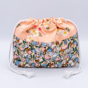 Pode incluir: Uma bolsa com cord&atilde;o apresentando uma se&ccedil;&atilde;o superior na cor p&ecirc;ssego com um padr&atilde;o floral branco e amarelo e uma se&ccedil;&atilde;o inferior com uma estampa bot&acirc;nica densa e multicolorida em tons de azul, rosa, amarelo e marrom, com cord&otilde;es brancos.