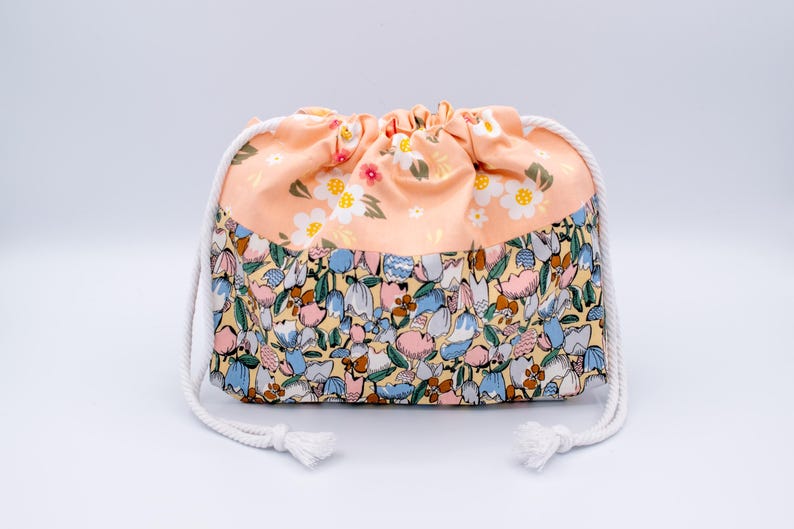 Pode incluir: Uma bolsa com cord&atilde;o apresentando uma se&ccedil;&atilde;o superior na cor p&ecirc;ssego com um padr&atilde;o floral branco e amarelo e uma se&ccedil;&atilde;o inferior com uma estampa bot&acirc;nica densa e multicolorida em tons de azul, rosa, amarelo e marrom, com cord&otilde;es brancos.