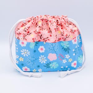 May include: A fabric drawstring pouch featuring a two-tone design. The upper section is light pink with a small red berry and branch pattern, while the lower section is light blue with a multi-colored floral print. It has white rope drawstrings.