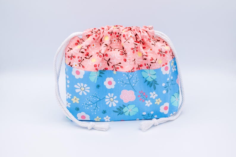 May include: A fabric drawstring pouch featuring a two-tone design. The upper section is light pink with a small red berry and branch pattern, while the lower section is light blue with a multi-colored floral print. It has white rope drawstrings.
