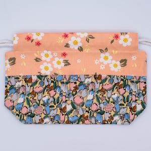 Pode incluir: Uma bolsa retangular de tecido com cord&atilde;o e design bicolor. A parte superior &eacute; cor de p&ecirc;ssego com um padr&atilde;o floral branco, enquanto a parte inferior exibe uma estampa densa e multicolorida de tulipas estilizadas em azul, rosa e amarelo.