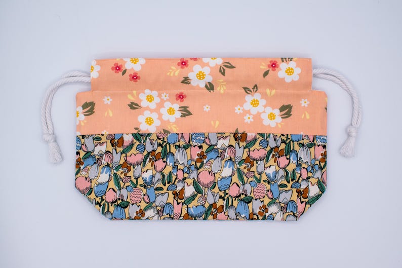 Pode incluir: Uma bolsa retangular de tecido com cord&atilde;o e design bicolor. A parte superior &eacute; cor de p&ecirc;ssego com um padr&atilde;o floral branco, enquanto a parte inferior exibe uma estampa densa e multicolorida de tulipas estilizadas em azul, rosa e amarelo.