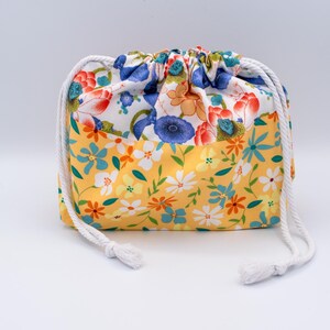 Puede incluir: Una bolsa de tela con cord&oacute;n ajustable y un patr&oacute;n floral de dos tonos. La secci&oacute;n superior muestra un fondo blanco con flores azules, naranjas y rojas, mientras que la secci&oacute;n inferior tiene un fondo amarillo con flores blancas, azules y naranjas.