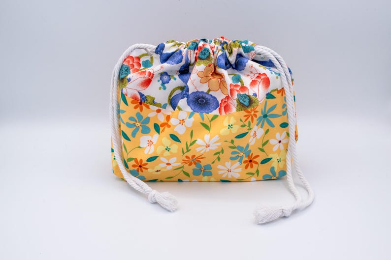 Puede incluir: Una bolsa de tela con cord&oacute;n ajustable y un patr&oacute;n floral de dos tonos. La secci&oacute;n superior muestra un fondo blanco con flores azules, naranjas y rojas, mientras que la secci&oacute;n inferior tiene un fondo amarillo con flores blancas, azules y naranjas.