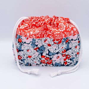 May include: A drawstring pouch featuring a two-tone floral pattern. The top section is a vibrant orange with white floral details, while the bottom section displays a dense mix of pink, red, blue, and white flowers. It has thick white rope cords.