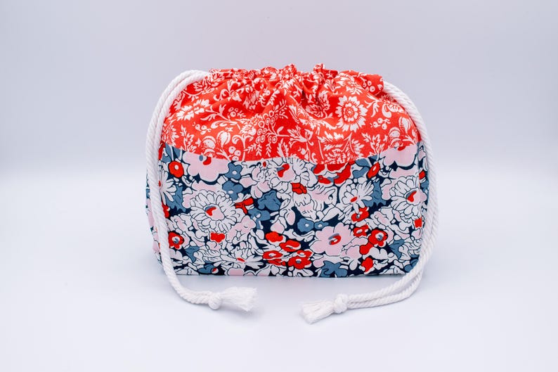 May include: A drawstring pouch featuring a two-tone floral pattern. The top section is a vibrant orange with white floral details, while the bottom section displays a dense mix of pink, red, blue, and white flowers. It has thick white rope cords.