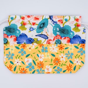 Puede incluir: Una bolsa rectangular de tela con cord&oacute;n, que presenta un dise&ntilde;o de dos partes. La secci&oacute;n superior muestra un fondo blanco con hojas de loto azules y verde azulado y flores de loto rojas. La secci&oacute;n inferior tiene un fondo amarillo con peque&ntilde;as flores naranjas, blancas y azules.