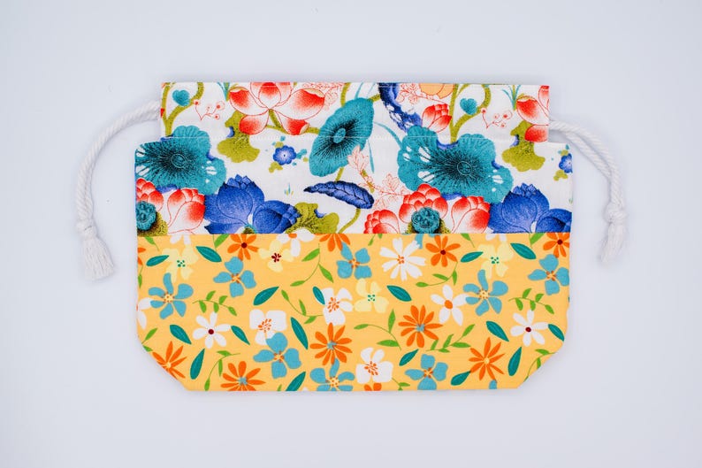 Puede incluir: Una bolsa rectangular de tela con cord&oacute;n, que presenta un dise&ntilde;o de dos partes. La secci&oacute;n superior muestra un fondo blanco con hojas de loto azules y verde azulado y flores de loto rojas. La secci&oacute;n inferior tiene un fondo amarillo con peque&ntilde;as flores naranjas, blancas y azules.