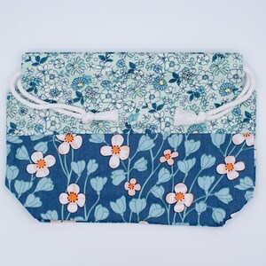 May include: A rectangular fabric drawstring pouch featuring two distinct floral patterns. The top section displays a dense light blue and white small flower print, while the bottom section features larger white flowers with orange centers on a dark blue background.