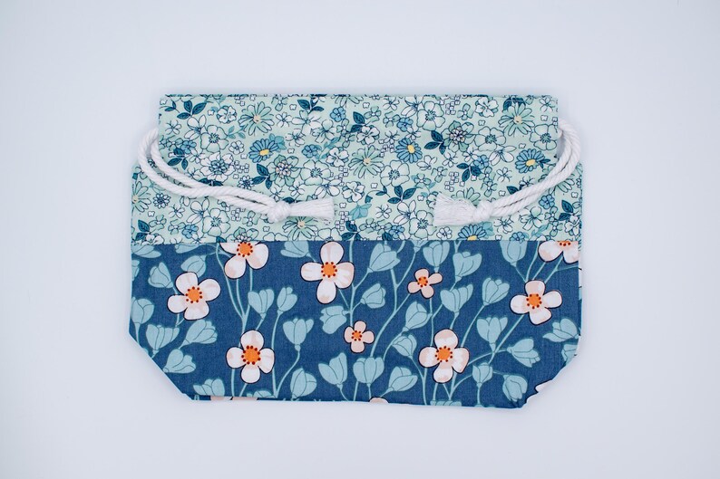 May include: A rectangular fabric drawstring pouch featuring two distinct floral patterns. The top section displays a dense light blue and white small flower print, while the bottom section features larger white flowers with orange centers on a dark blue background.