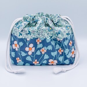 May include: A fabric drawstring pouch featuring a two-tone floral pattern. The upper section has a light blue background with small white and blue flowers, while the lower section is a darker blue with larger white flowers with orange centers.