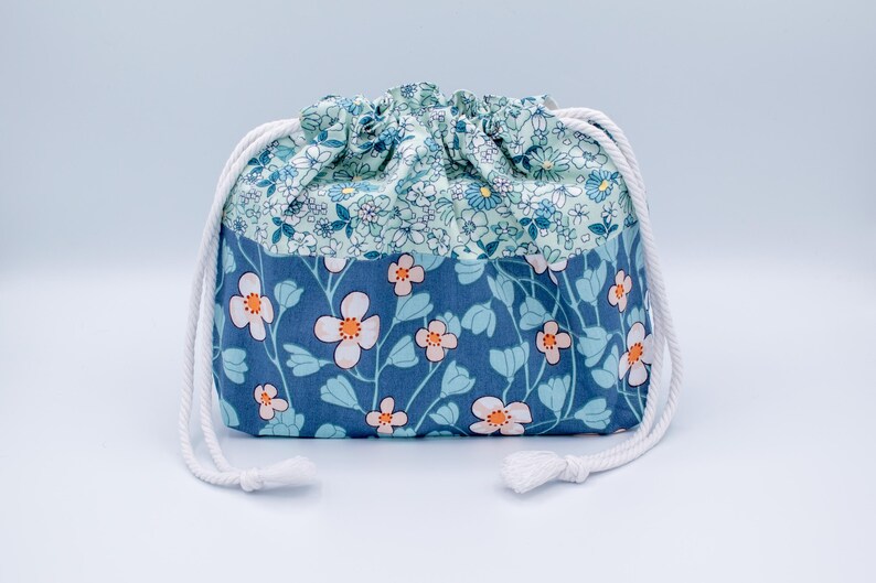 May include: A fabric drawstring pouch featuring a two-tone floral pattern. The upper section has a light blue background with small white and blue flowers, while the lower section is a darker blue with larger white flowers with orange centers.