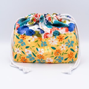 Puede incluir: Una bolsa de tela con cord&oacute;n ajustable y un patr&oacute;n floral de dos tonos. La secci&oacute;n inferior es amarilla con peque&ntilde;as flores blancas, naranjas y azules, mientras que la secci&oacute;n superior muestra motivos florales m&aacute;s grandes en azul y rojo.
