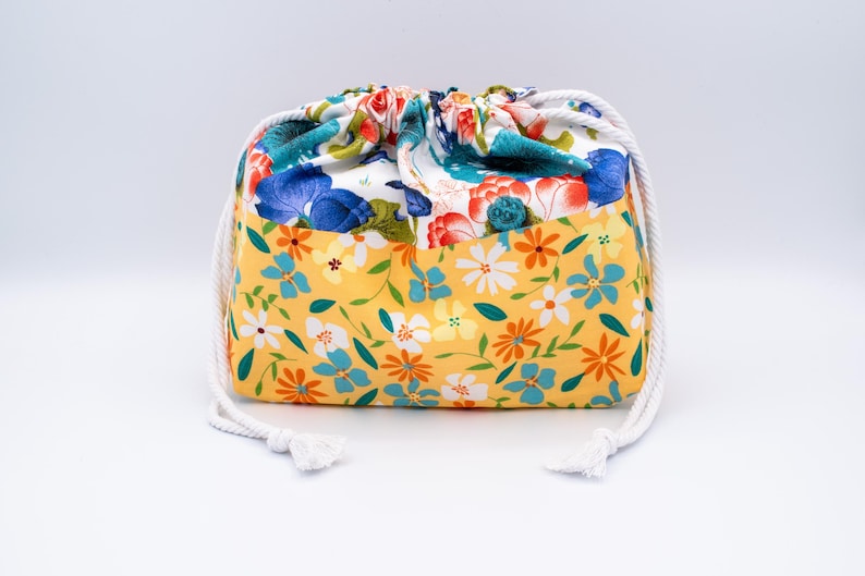 Puede incluir: Una bolsa de tela con cord&oacute;n ajustable y un patr&oacute;n floral de dos tonos. La secci&oacute;n inferior es amarilla con peque&ntilde;as flores blancas, naranjas y azules, mientras que la secci&oacute;n superior muestra motivos florales m&aacute;s grandes en azul y rojo.