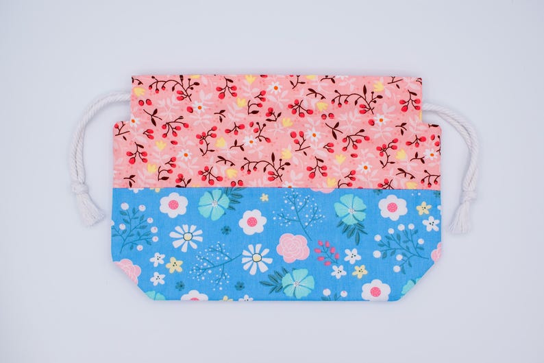 May include: A rectangular drawstring pouch featuring a two-tone design. The top section is a light coral fabric with a pattern of small white flowers and dark red berries. The bottom section is a bright blue fabric with white and pink flowers.