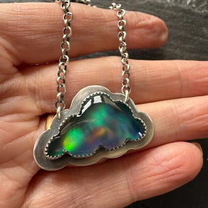 May include: A silver cloud-shaped pendant necklace with a colorful, iridescent center. The pendant has a textured border and hangs from a delicate chain. The iridescent center displays shades of green, blue, and yellow.