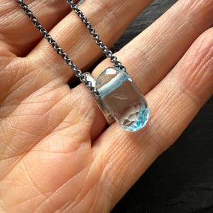 May include: A clear, faceted pendant necklace with a light blue gemstone. The pendant is suspended from a dark silver chain. The necklace is displayed on a person's open palm.