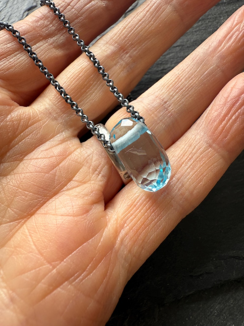 May include: A clear, faceted pendant necklace with a light blue gemstone. The pendant is suspended from a dark silver chain. The necklace is displayed on a person's open palm.