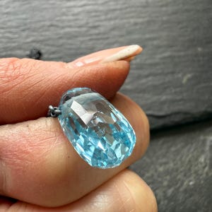 May include: A faceted, light blue gemstone pendant. The pendant is a large, elongated teardrop shape with many facets that catch the light. The gemstone is suspended from a delicate silver chain. The background is a dark, textured surface.