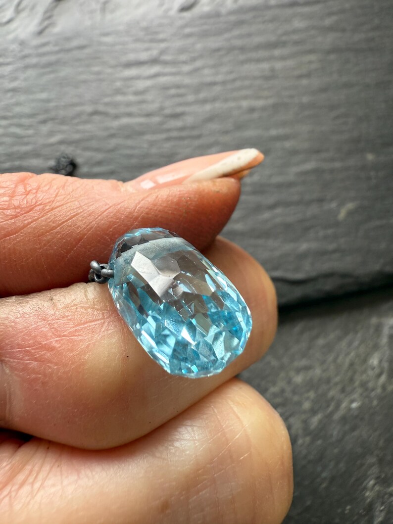 May include: A faceted, light blue gemstone pendant. The pendant is a large, elongated teardrop shape with many facets that catch the light. The gemstone is suspended from a delicate silver chain. The background is a dark, textured surface.