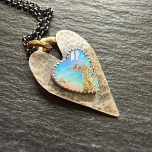May include: A heart-shaped pendant necklace with a silver chain. The pendant features a hammered silver heart with a smaller, textured heart-shaped opal stone in the centre. The opal displays blue, green, and orange hues.