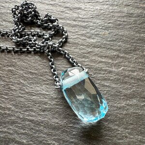 May include: A faceted, light blue gemstone pendant necklace. The pendant is a teardrop shape, suspended from a dark, textured chain. The necklace is displayed on a dark gray, textured surface.