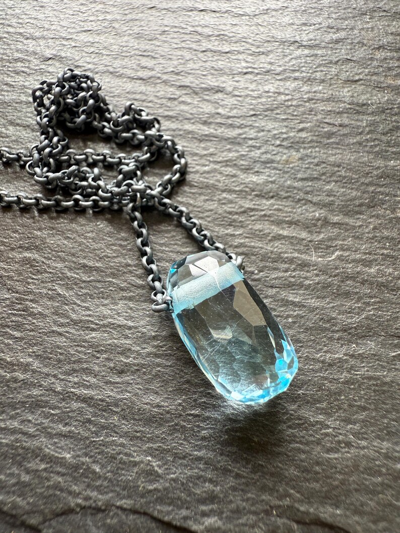 May include: A faceted, light blue gemstone pendant necklace. The pendant is a teardrop shape, suspended from a dark, textured chain. The necklace is displayed on a dark gray, textured surface.