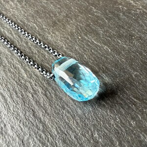 May include: A faceted, light blue gemstone pendant necklace. The pendant is a large, elongated teardrop shape with multiple facets. The chain is a dark, oxidized silver color. The necklace is displayed on a dark gray, textured surface.