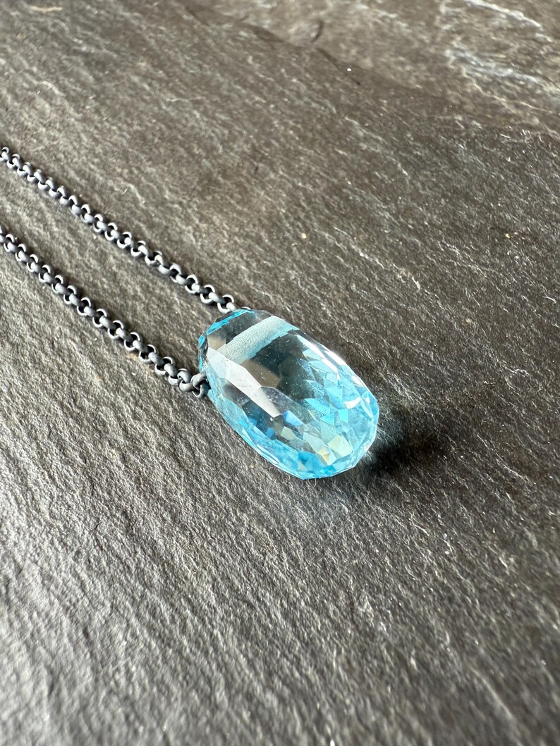 May include: A faceted, light blue gemstone pendant necklace. The pendant is a large, elongated teardrop shape with multiple facets. The chain is a dark, oxidized silver color. The necklace is displayed on a dark gray, textured surface.