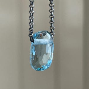 May include: A faceted, light blue gemstone pendant suspended from a dark silver chain. The gemstone is a rectangular shape with multiple facets, reflecting light. The chain is a series of small, linked rings.