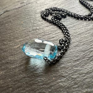 May include: A faceted, teardrop-shaped, light blue gemstone pendant necklace. The pendant is suspended from a dark, oxidized chain. The necklace is displayed on a dark, textured surface. Jewelry.