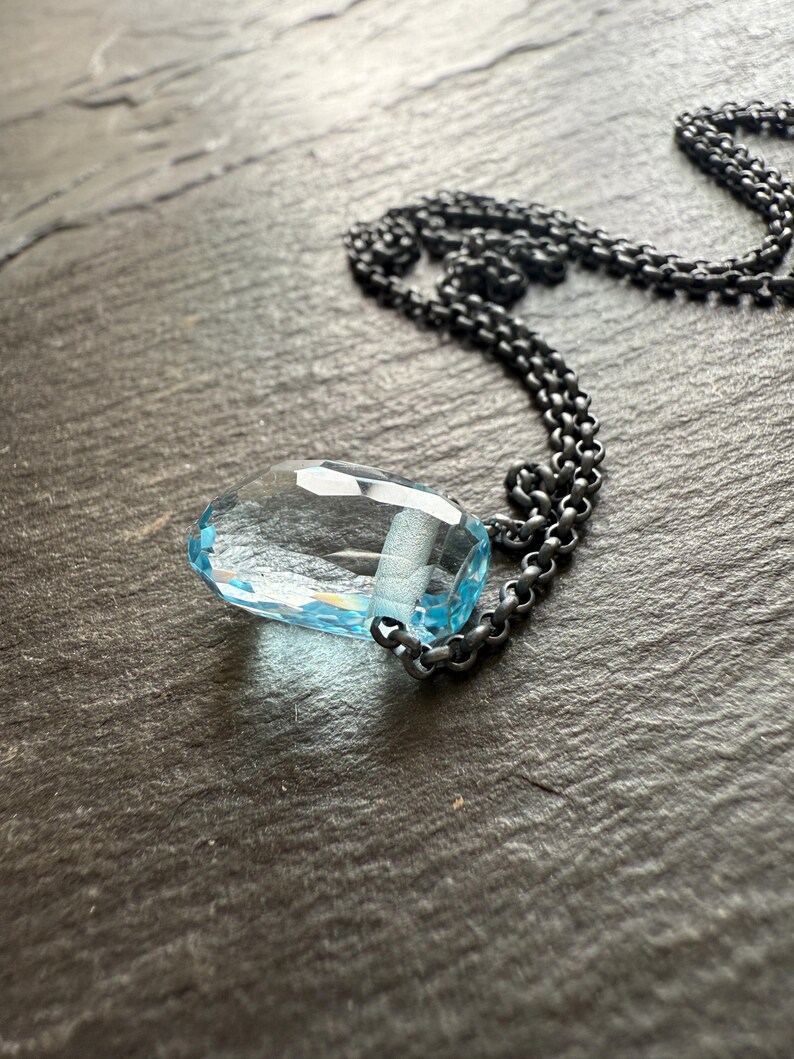 May include: A faceted, teardrop-shaped, light blue gemstone pendant necklace. The pendant is suspended from a dark, oxidized chain. The necklace is displayed on a dark, textured surface. Jewelry.