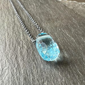 May include: A silver chain necklace with a large, faceted, light blue gemstone pendant. The pendant is teardrop-shaped and catches the light, showcasing its intricate cut. The chain is a delicate, beaded design.