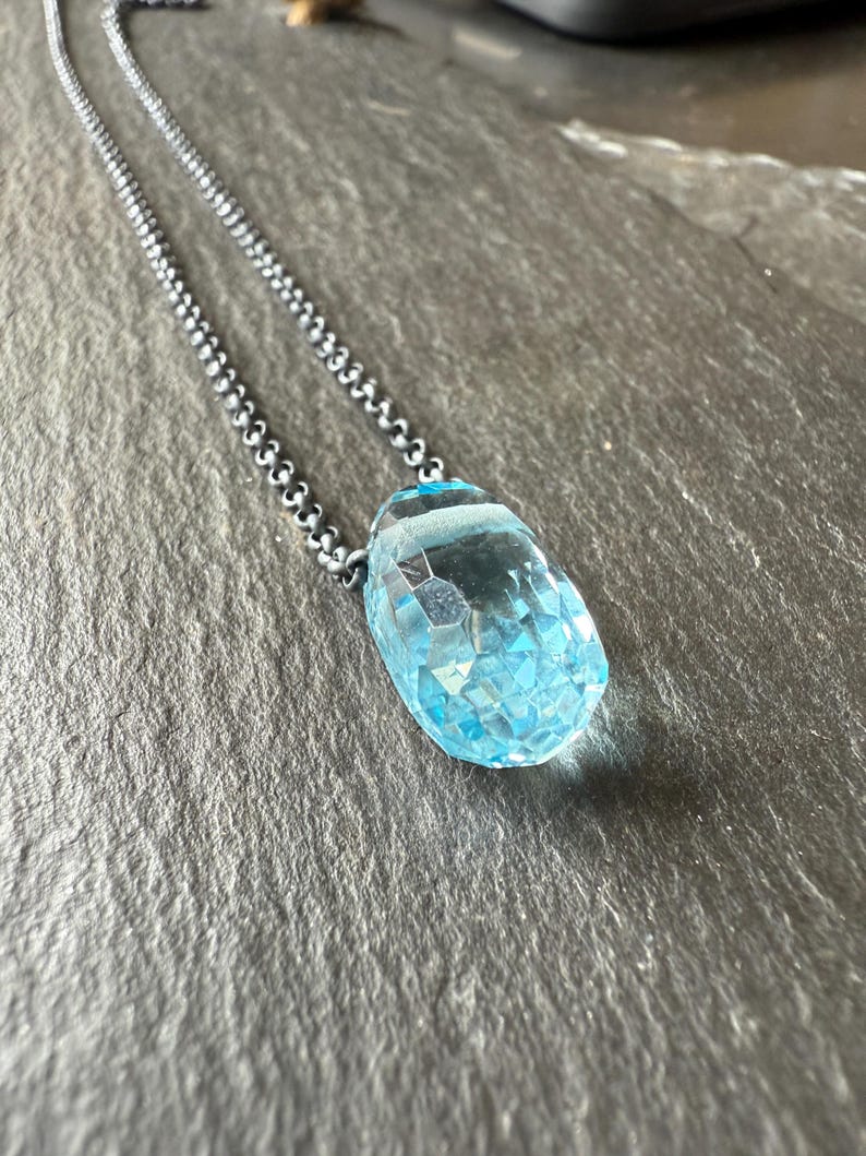 May include: A silver chain necklace with a large, faceted, light blue gemstone pendant. The pendant is teardrop-shaped and catches the light, showcasing its intricate cut. The chain is a delicate, beaded design.