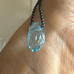 May include: A faceted, light blue gemstone pendant necklace. The pendant is a teardrop shape and is suspended from a dark, beaded chain. The gemstone has a clear, crystalline appearance.