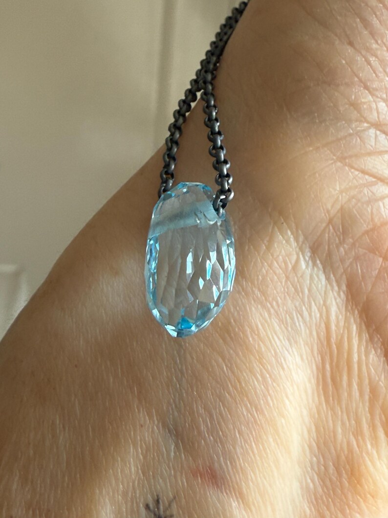 May include: A faceted, light blue gemstone pendant necklace. The pendant is a teardrop shape and is suspended from a dark, beaded chain. The gemstone has a clear, crystalline appearance.
