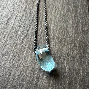 May include: A faceted, light blue gemstone pendant necklace. The pendant is a teardrop shape and is suspended from a delicate, oxidized silver chain. The necklace is displayed against a dark, textured background.
