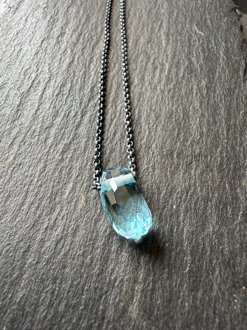 May include: A faceted, light blue gemstone pendant necklace. The pendant is a teardrop shape and is suspended from a delicate, oxidized silver chain. The necklace is displayed against a dark, textured background.