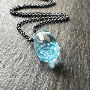 May include: A faceted, light blue gemstone pendant necklace with a dark, gunmetal-colored chain. The pendant is a large, teardrop shape. The chain is a linked design. The necklace is displayed on a dark, textured surface.