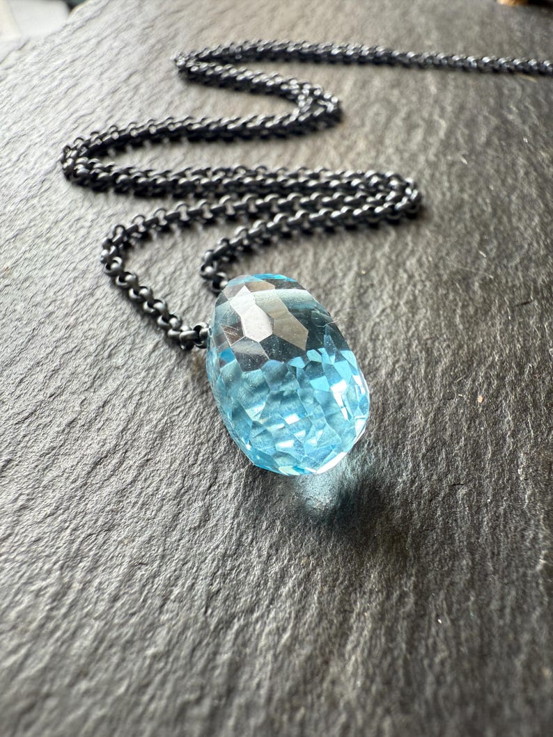 May include: A faceted, light blue gemstone pendant necklace with a dark, gunmetal-colored chain. The pendant is a large, teardrop shape. The chain is a linked design. The necklace is displayed on a dark, textured surface.
