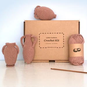 以下が含まれることがあります： Little Conkers Crochet Kitと書かれた茶色の段ボール箱に入ったかぎ針編みキット。くすんだピンク色のかぎ針編みアンフォラ花瓶3つ、どんぐり2つが描かれたラベル付きの毛糸玉、木製のかぎ針1本が含まれています。