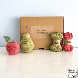 May include: A brown cardboard box labeled Little Conkers Crochet Kit, accompanied by a crocheted pink apple with a green leaf, a crocheted green pear, and two skeins of yarn in light green and pink, each wrapped with a paper band.