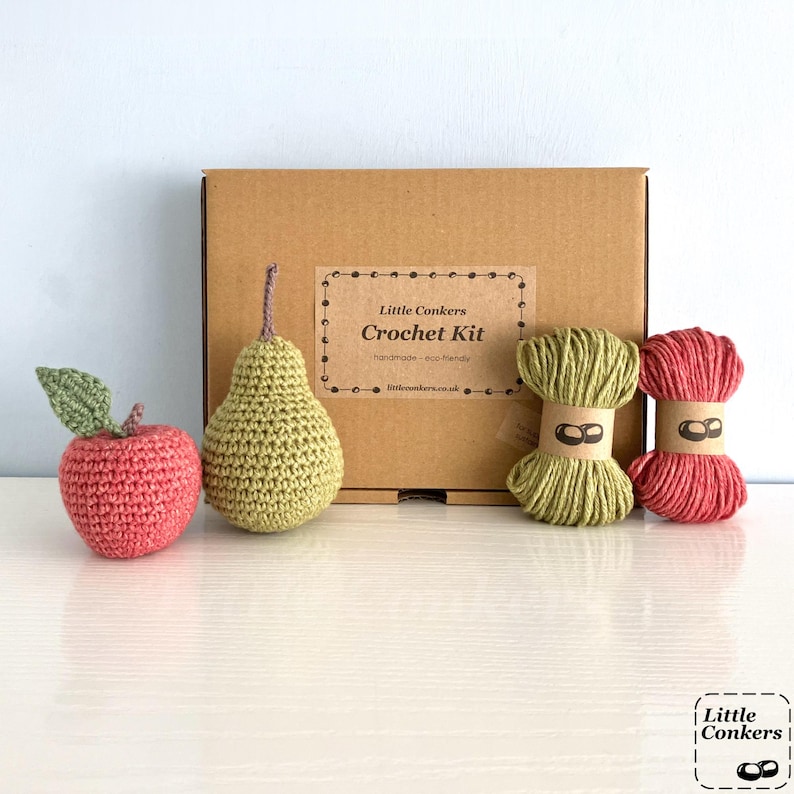 May include: A brown cardboard box labeled Little Conkers Crochet Kit, accompanied by a crocheted pink apple with a green leaf, a crocheted green pear, and two skeins of yarn in light green and pink, each wrapped with a paper band.