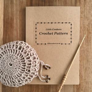 May include: A tan crochet pattern booklet titled Little Conkers Crochet Pattern with a website address, a round beige crocheted piece with dangling beads, and a light-colored wooden crochet hook with a decorative handle and a tapered tip.