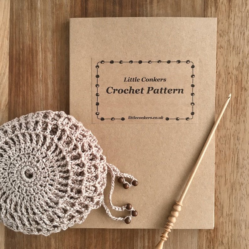 May include: A tan crochet pattern booklet titled Little Conkers Crochet Pattern with a website address, a round beige crocheted piece with dangling beads, and a light-colored wooden crochet hook with a decorative handle and a tapered tip.