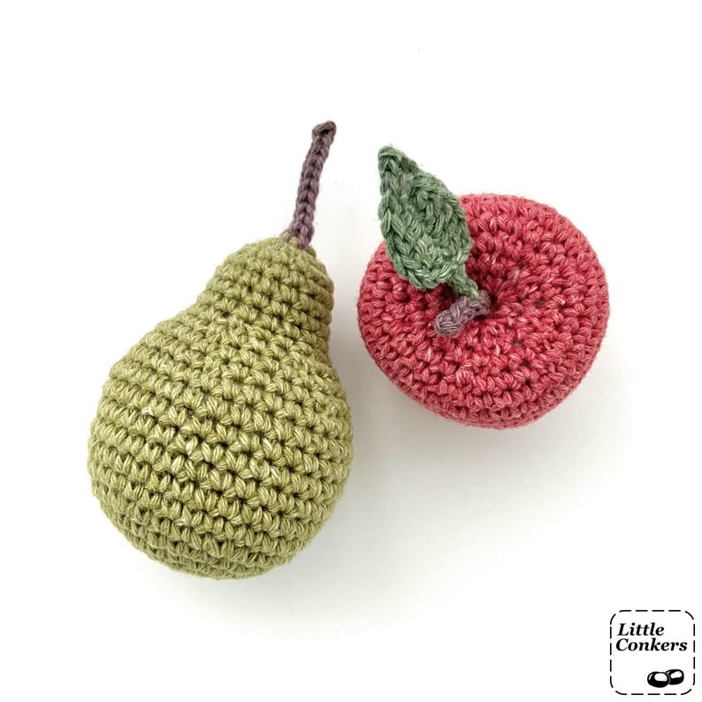 May include: A handmade crochet pear in olive green with a brown stem and a crochet apple in deep pink with a green leaf. The items feature a textured yarn stitch pattern. A small logo in the corner displays the text Little Conkers.