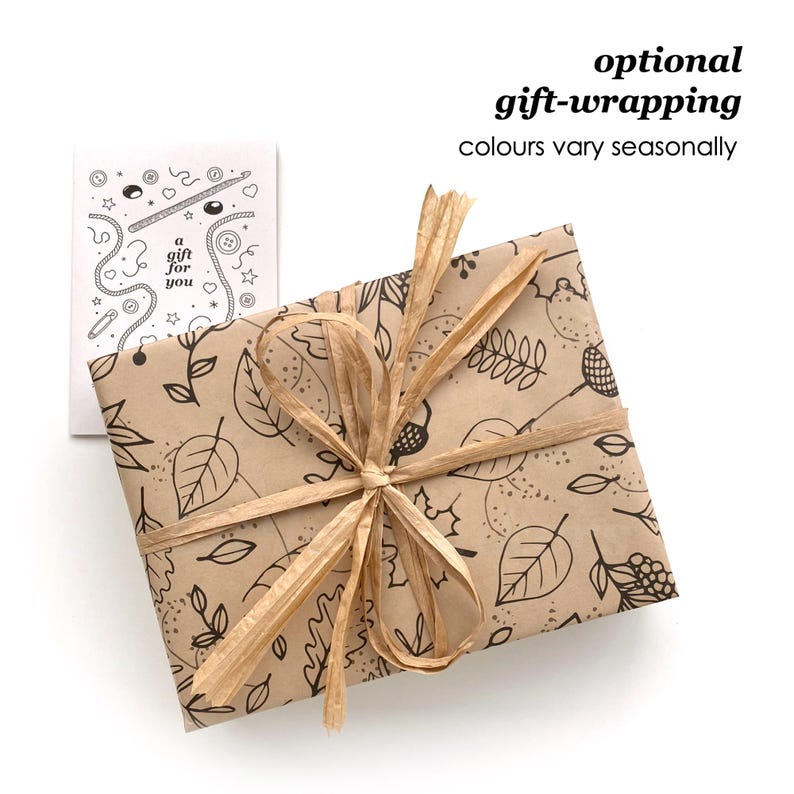 May include: A rectangular gift wrapped in brown paper with a black line-art pattern of leaves and acorns, tied with a tan raffia ribbon. Beside it is a white card with black line-art illustrations and the text 'a gift for you'.