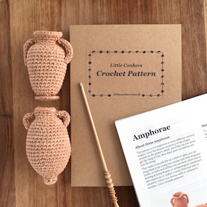 Könnte beinhalten: Häkelanleitung für die Herstellung von Miniatur-Amphoren, einer Art antiker griechischer Vase. Das Muster ist auf einem braunen Papierheft mit einer Häkelnadel und zwei fertigen Amphoren in hellpfirsichfarben gedruckt.